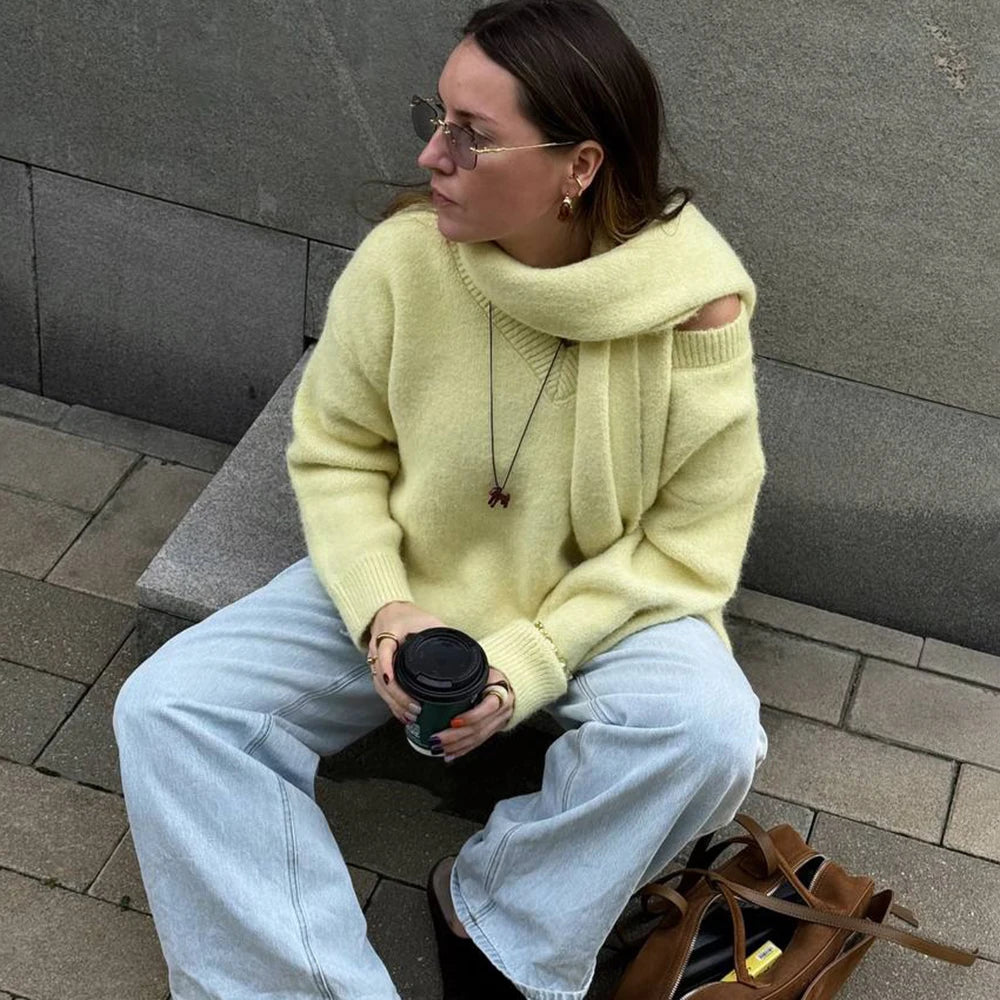 Woman in a light green sweater and blue jeans sitting on a bench holding a coffee cup.