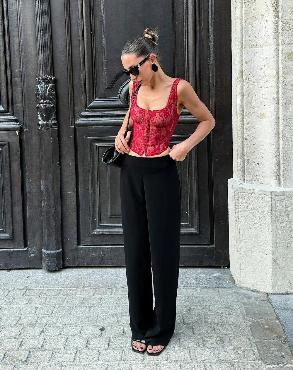 Woman in red lace top and black pants standing in front of a dark door.