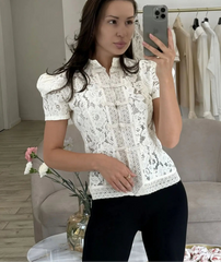 Woman taking a mirror selfie wearing a white lace blouse in a room with a gray armchair and clothing rack.