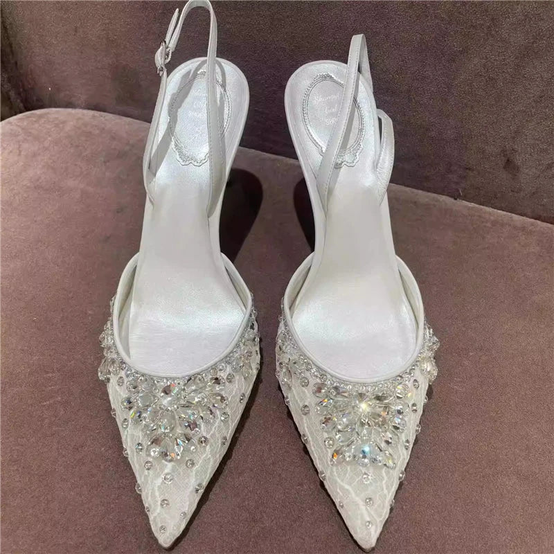 Pair of white high-heeled shoes with crystal embellishments on a brown surface.