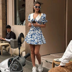 Woman in a blue floral dress standing on a street with a building and scooter in the background.