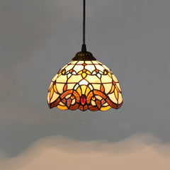 Golden Atelier pendant light featuring a handcrafted Tiffany glass shade with colorful stained glass panels, casting a warm glow. The light hangs from a chain on a ceiling in a living room setting. A