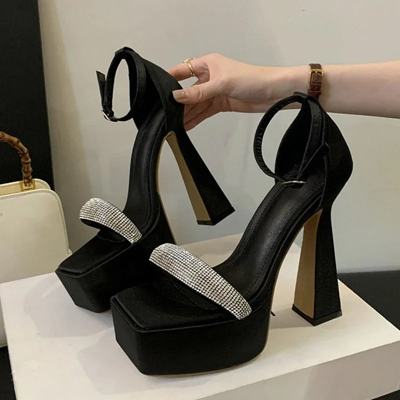 Black high-heeled shoes with rhinestone straps held by a hand on a white surface.