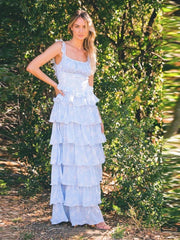 Woman wearing a light blue tiered dress standing in front of green foliage.