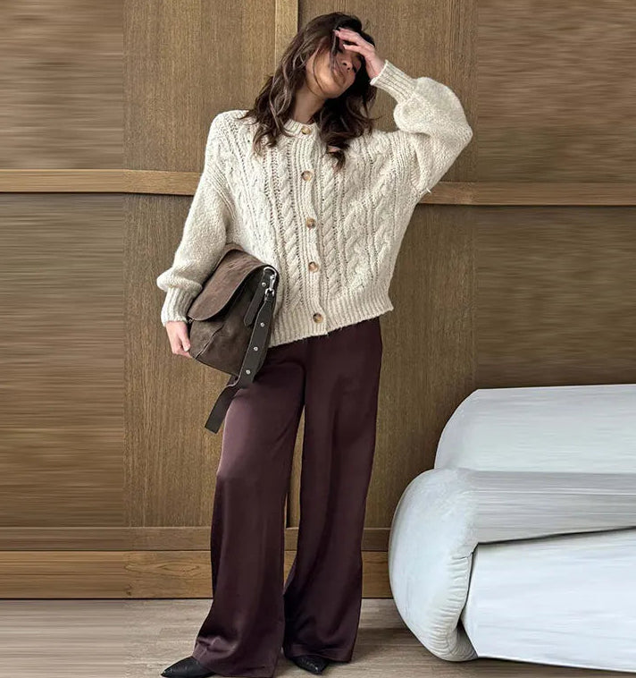 Woman wearing a cream sweater and brown pants holding a brown bag against a wooden wall.