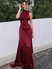 Woman wearing a burgundy evening gown standing outdoors against a white wall with greenery.