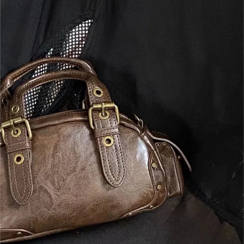 Brown leather handbag with gold buckles held by a person wearing a black outfit.