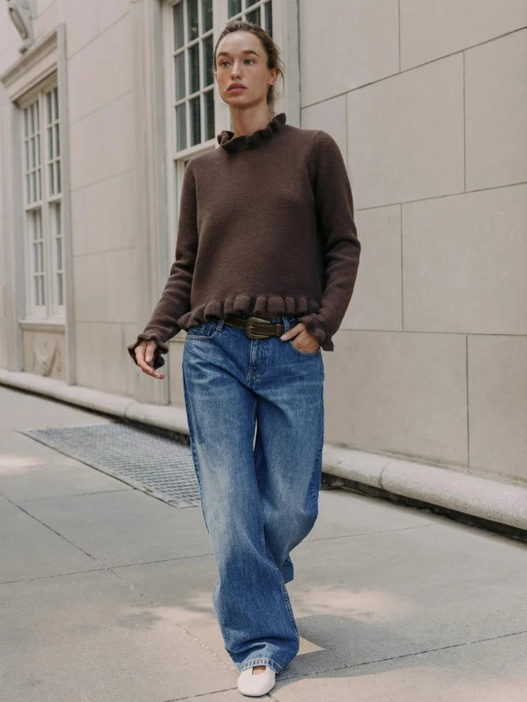 Person wearing a brown sweater and blue jeans standing on a sidewalk.