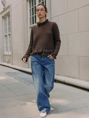 Person wearing a brown sweater and blue jeans standing on a sidewalk.