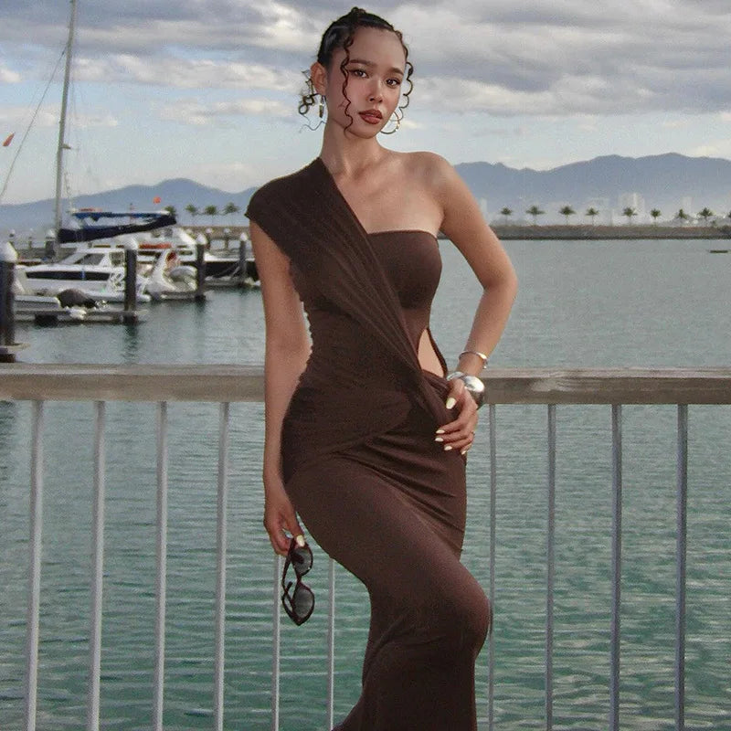 Woman in a brown one-shoulder dress standing by a waterfront with boats and mountains in the background.