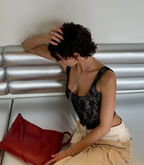 Person sitting on a metallic bench holding a red bag, wearing a black lace top and beige pants.