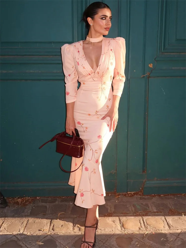 Woman in a floral dress holding a brown handbag against a teal wall.