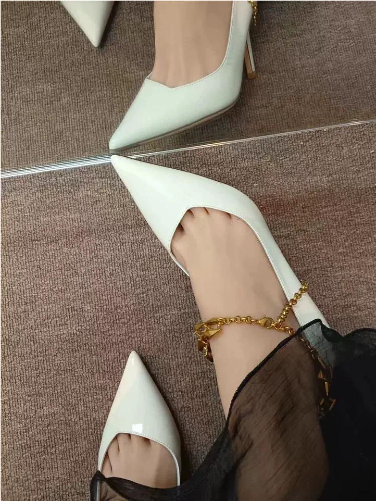 White high-heeled shoes worn with a gold anklet on a brown floor.