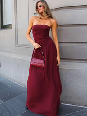Woman in a burgundy strapless gown holding a matching handbag against a neutral wall.