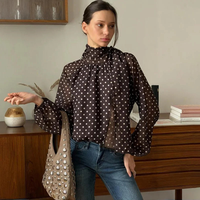 Woman wearing a brown polka dot blouse and blue jeans, holding a beige handbag in a room with wooden furniture.