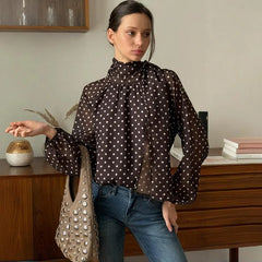 Woman wearing a brown polka dot blouse and blue jeans, holding a beige handbag in a room with wooden furniture.