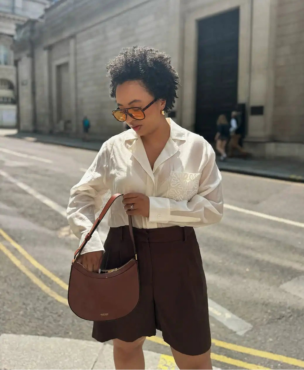 Woman walking on a street wearing a white blouse and brown shorts, holding a brown handbag.