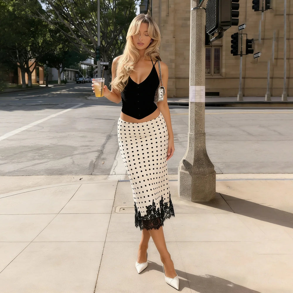 Woman in black top and white polka dot skirt standing on a city street holding a drink.