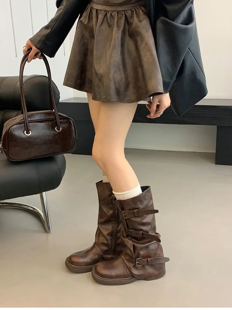 Person wearing brown leather boots, holding a brown handbag indoors.