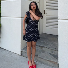 Woman in a black polka dot dress with red shoes standing on steps outside a building.