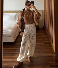 Woman taking a mirror selfie wearing a brown blouse and white lace pants in a bedroom.