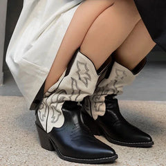 Black cowboy boots with decorative leg warmers worn by a person sitting on a concrete surface.