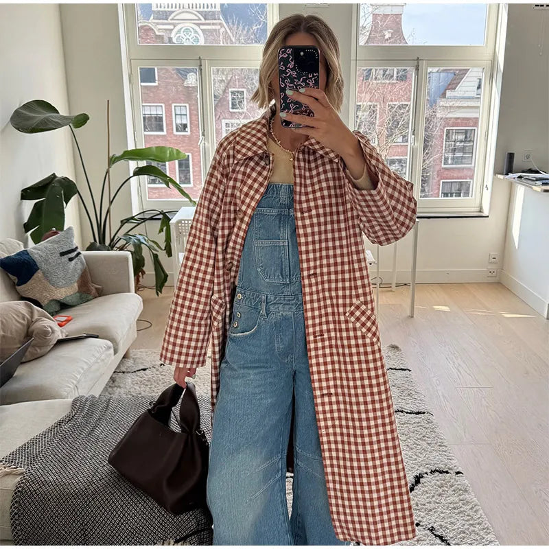 Person taking a mirror selfie wearing a checkered coat and denim overalls in a living room.