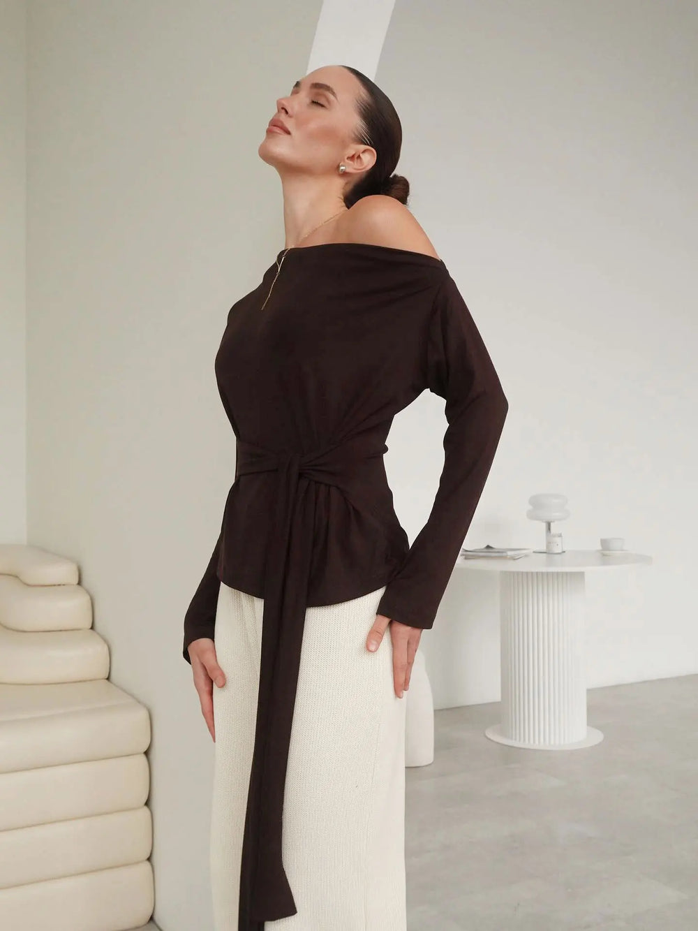 Woman wearing a black off-shoulder top and white pants in a minimalistic room.