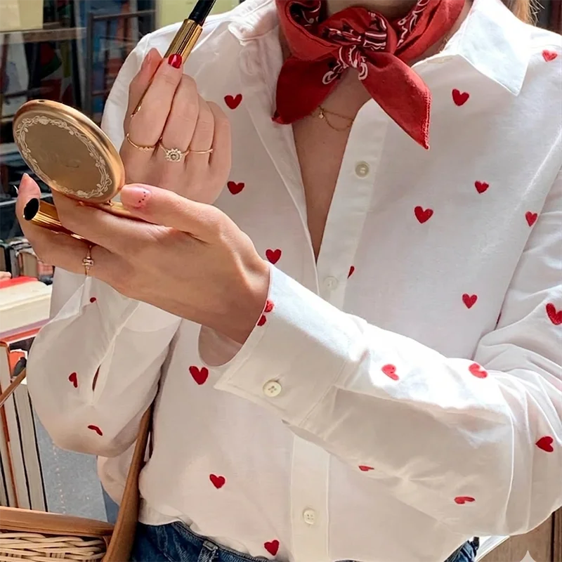Person wearing a white blouse with red heart patterns, holding a compact mirror.
