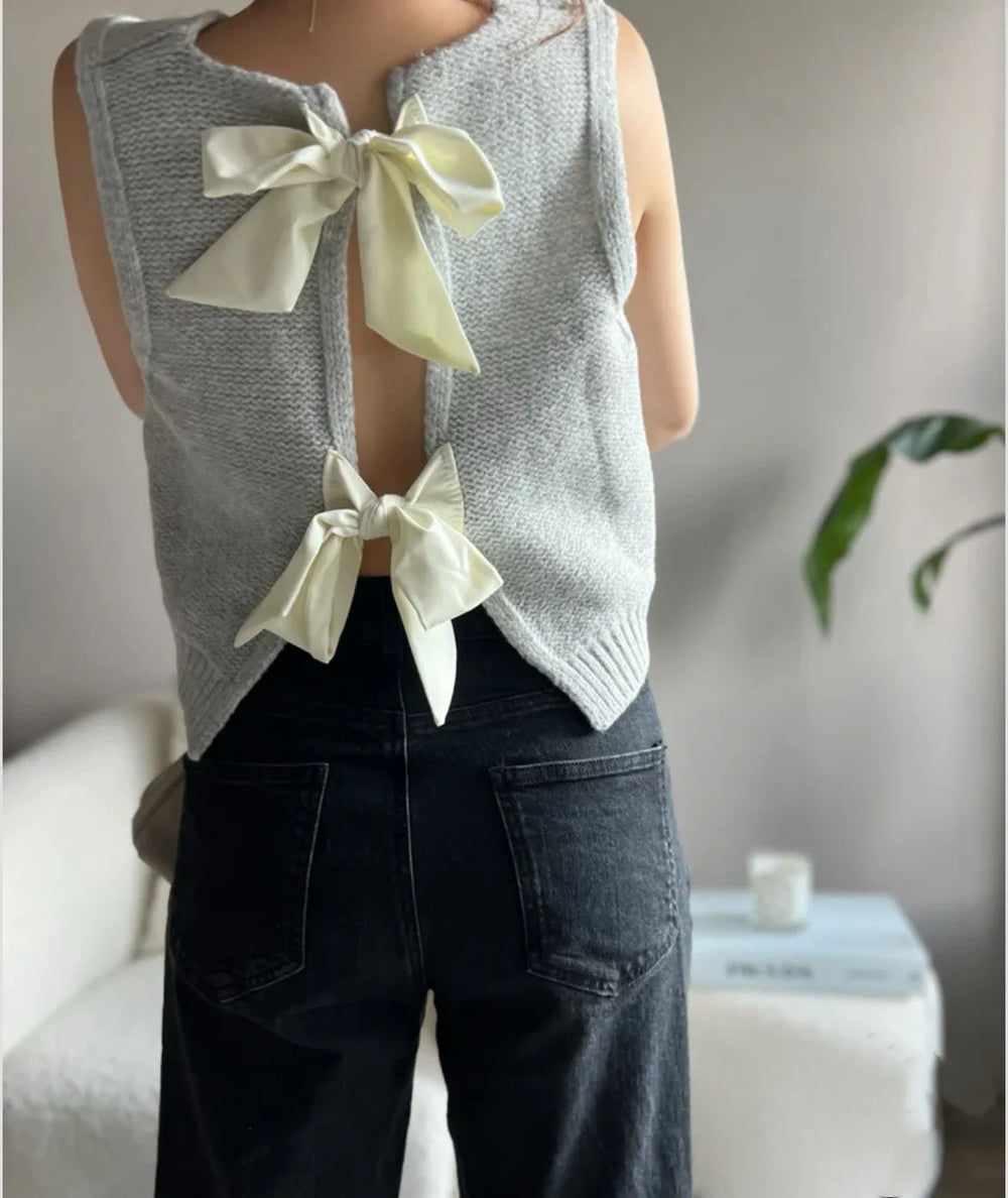 Person wearing a gray sleeveless top with white bows and dark jeans in an indoor setting.