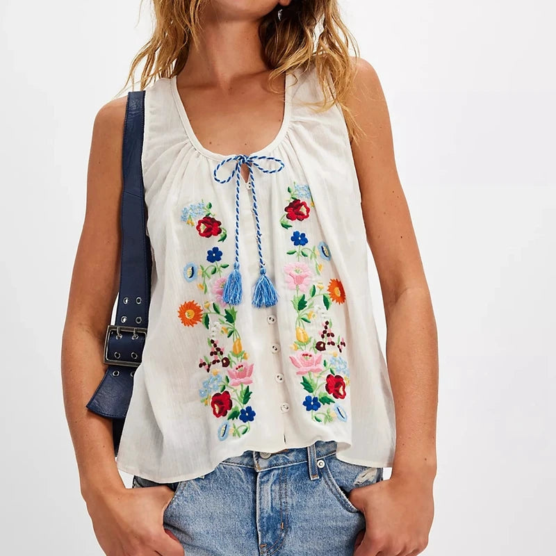 Woman wearing a white sleeveless top with colorful floral embroidery on a plain background
