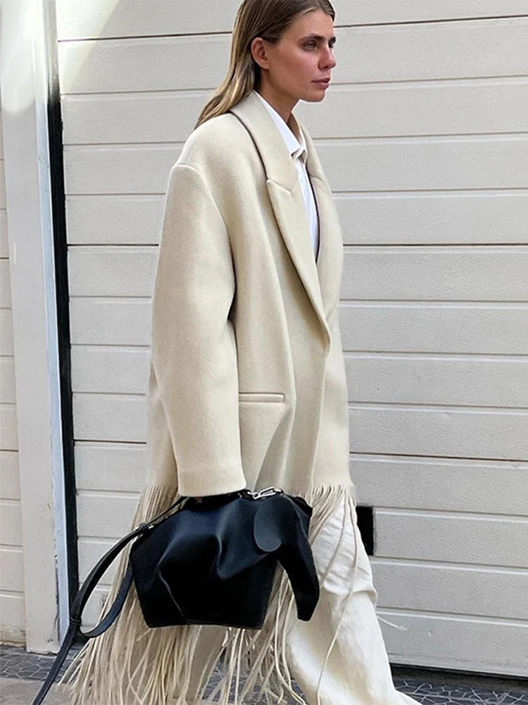 Woman wearing a long cream coat with a black handbag against a light-colored wall.