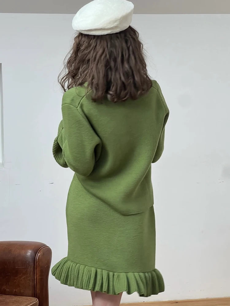 Person wearing a green dress with a white beret from behind, standing against a plain white wall.
