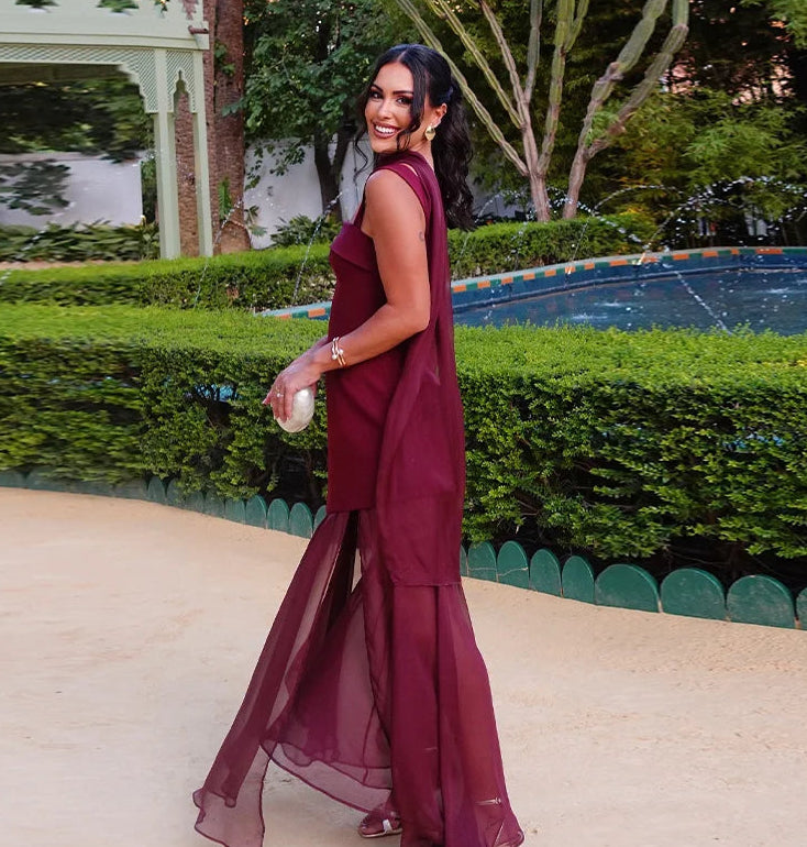 Woman in a burgundy dress standing outdoors with greenery and a pool in the background