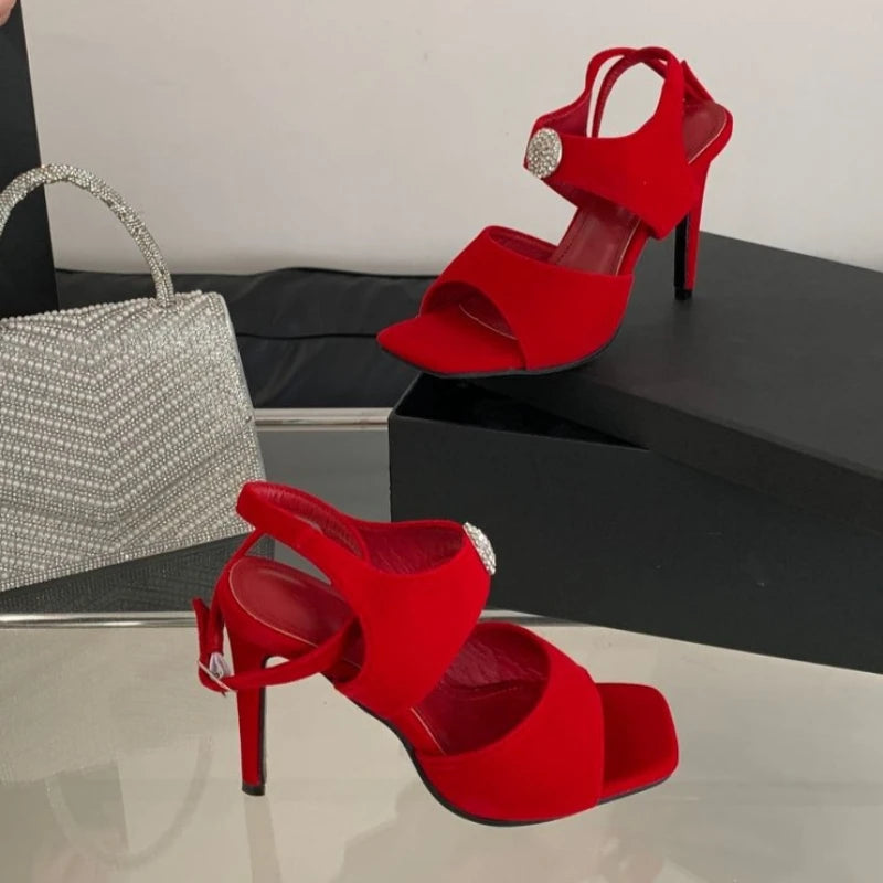 Red stiletto heels with rhinestone embellishment and ankle strap next to a silver handbag.