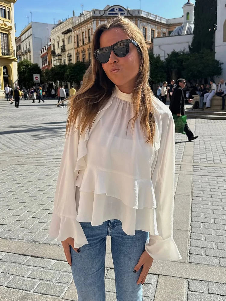 Woman wearing a white blouse and sunglasses in an outdoor setting with people and buildings in the background.