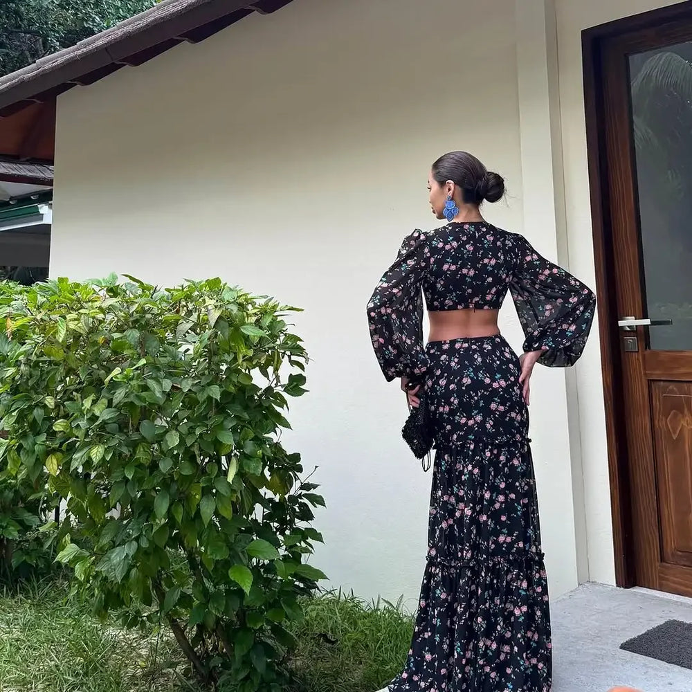 Woman in a floral dress standing outdoors next to a building with greenery.