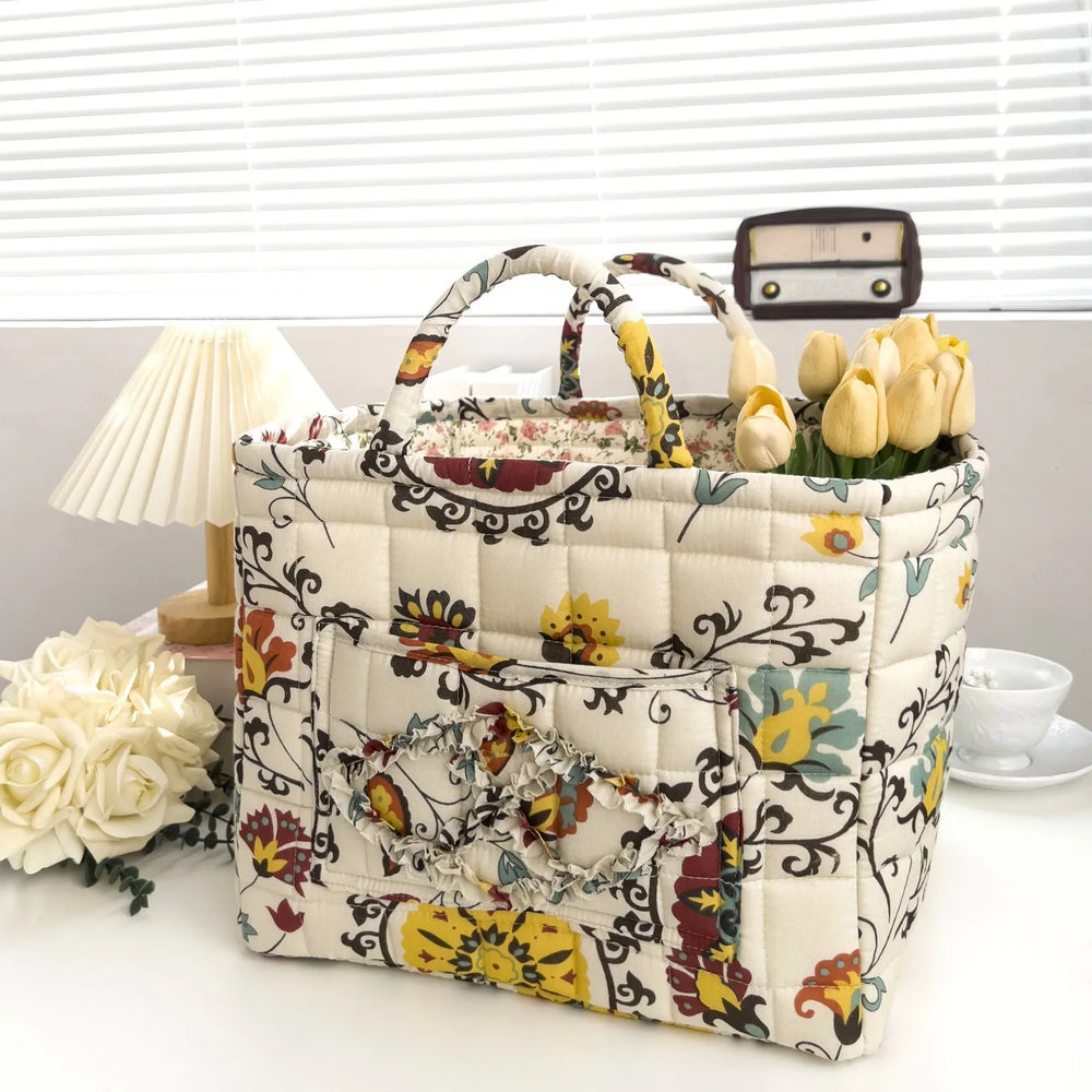 Floral-patterned quilted bag on a white surface with flowers and a lamp in the background