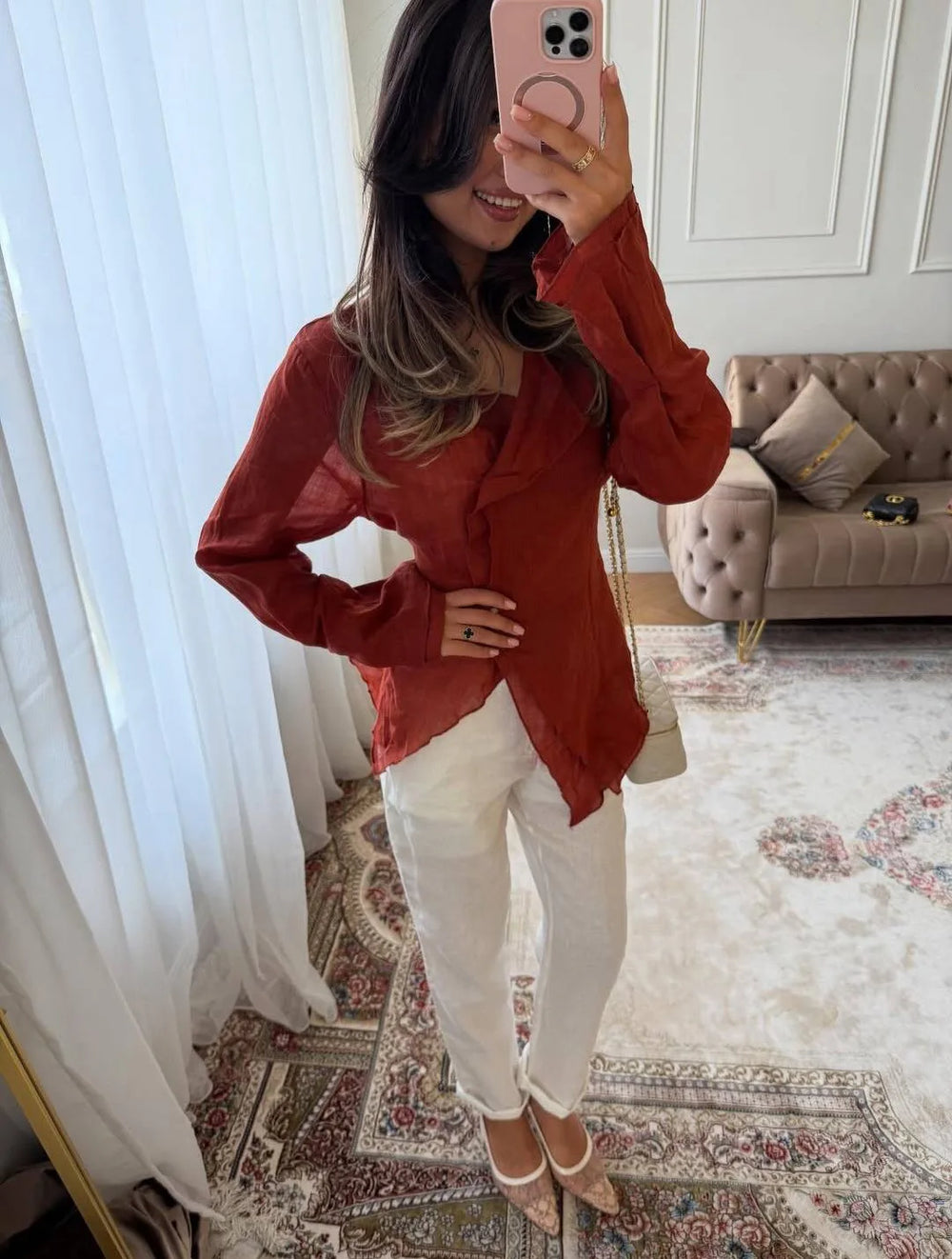 Woman taking a mirror selfie wearing a red blouse and white pants in a room with a sofa and curtains.