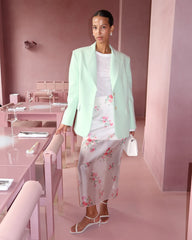 Woman in a light green blazer and floral dress standing in a pink room.