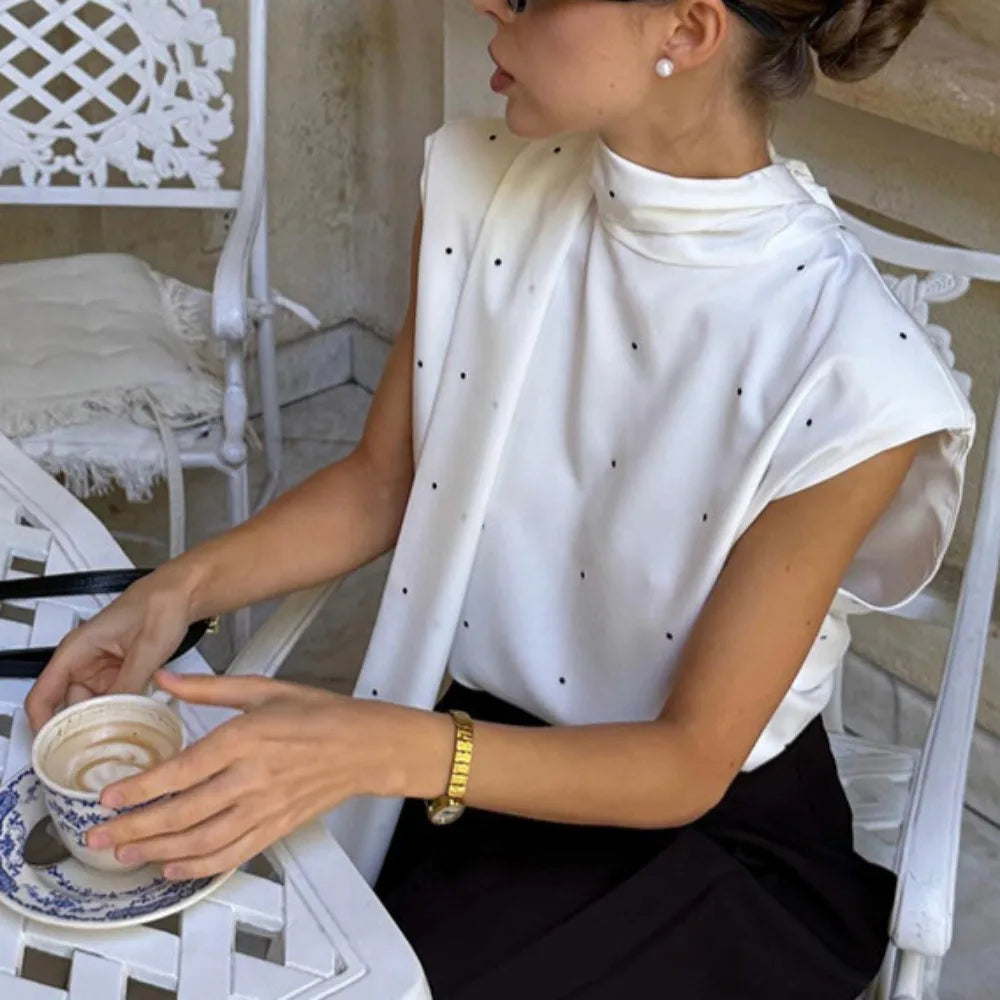 Woman sitting outdoors holding a cup of coffee, wearing a white blouse with black polka dots.