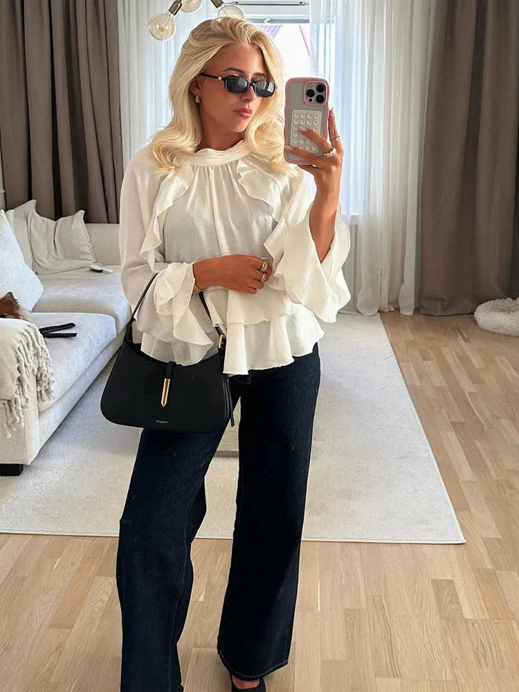 Woman taking a mirror selfie in a living room wearing a white blouse and black pants.