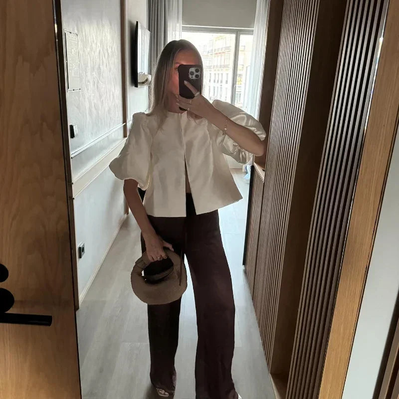 Person taking a mirror selfie wearing a white blouse and brown pants, holding a beige hat.