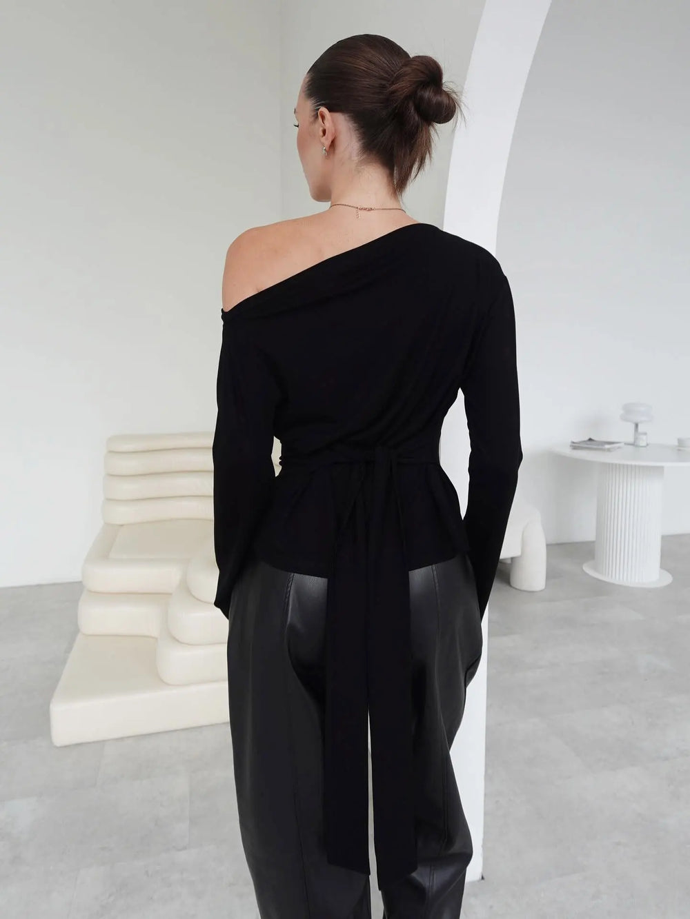Person wearing a black off-shoulder top and black pants in a minimalistic room.