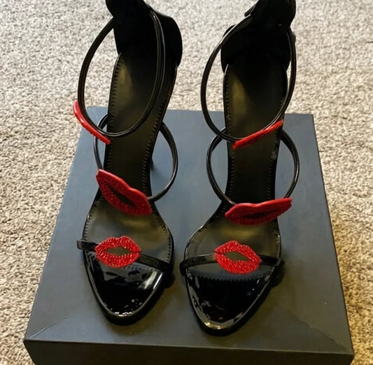 Pair of black patent leather stiletto sandals with red lip embellishments on a box