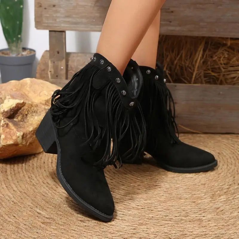 Black fringed boots worn by a person on a woven surface with a wooden bench and plant in the background.