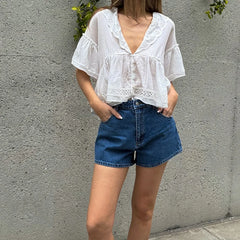 Person wearing a white blouse and blue shorts against a gray wall.