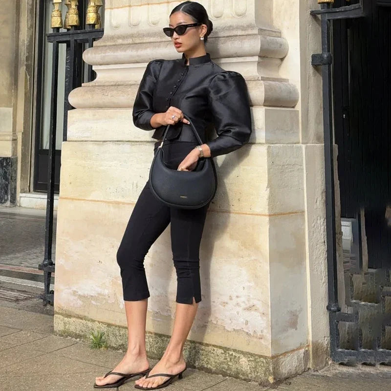 Woman in black outfit holding a matching handbag against a stone wall.