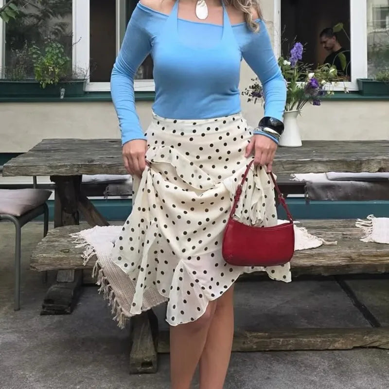 Woman in a blue top and polka dot skirt holding a red purse outdoors.