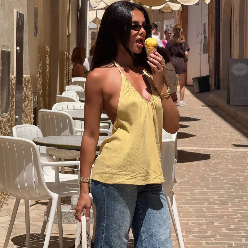 Woman in yellow top and blue jeans eating an ice cream on a street with tables and chairs.